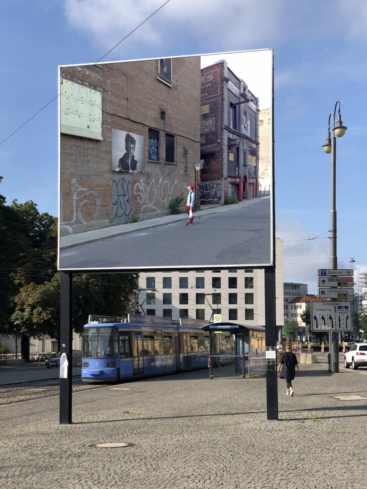 Ein riesiges Bild auf Stangen mitten in der Stadt, darauf ist ein Clown, der auf ein Poster an einer Hauswand schaut abgebildet